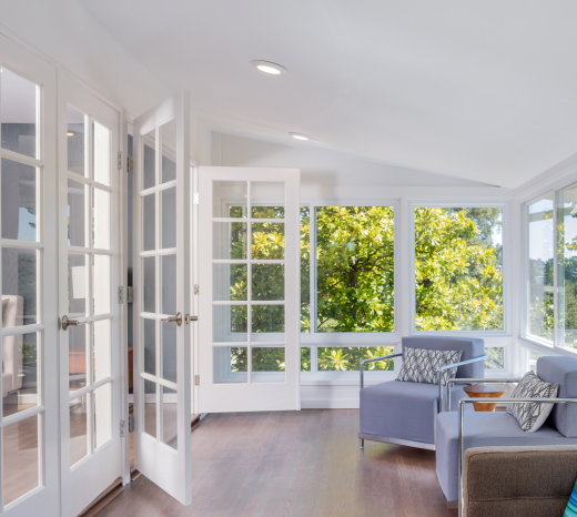 Modern sunroom with white ceilings, doors, and abundant greenery outside. LED bulbs illuminate the room, creating a bright and airy atmosphere.