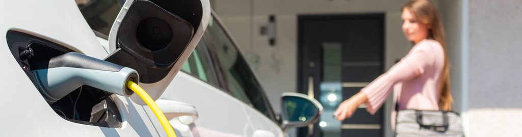A close up image of an EV charger plugged into an EV. The car owner is shown in the backround locking her car with her key fob.