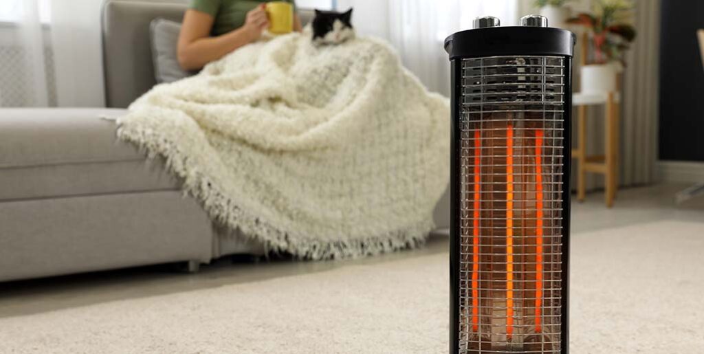 Woman with mug and cat, wrapped in blanket, sitting on couch, with space heater in foreground.